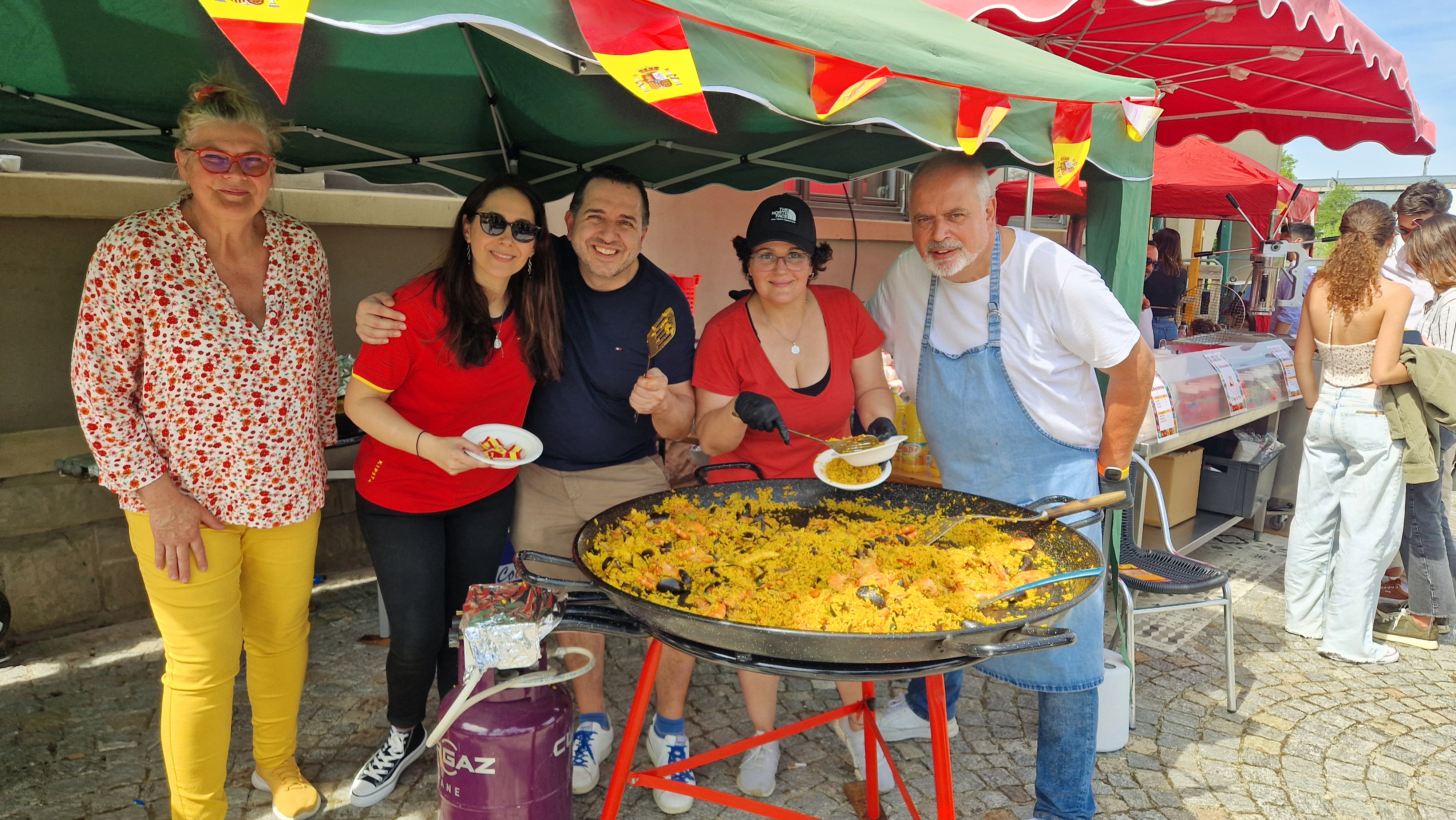 A group of five volunteers smiling and posing in front of a large paella dish at a festival.